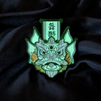 A green and white Chinese traditional lion head enamel pin, displayed on a dark black fabric background. the white paint glows green in the dark