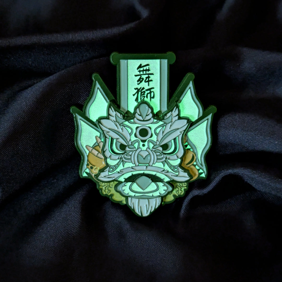 A green and white Chinese traditional lion head enamel pin, displayed on a dark black fabric background. the white paint glows green in the dark